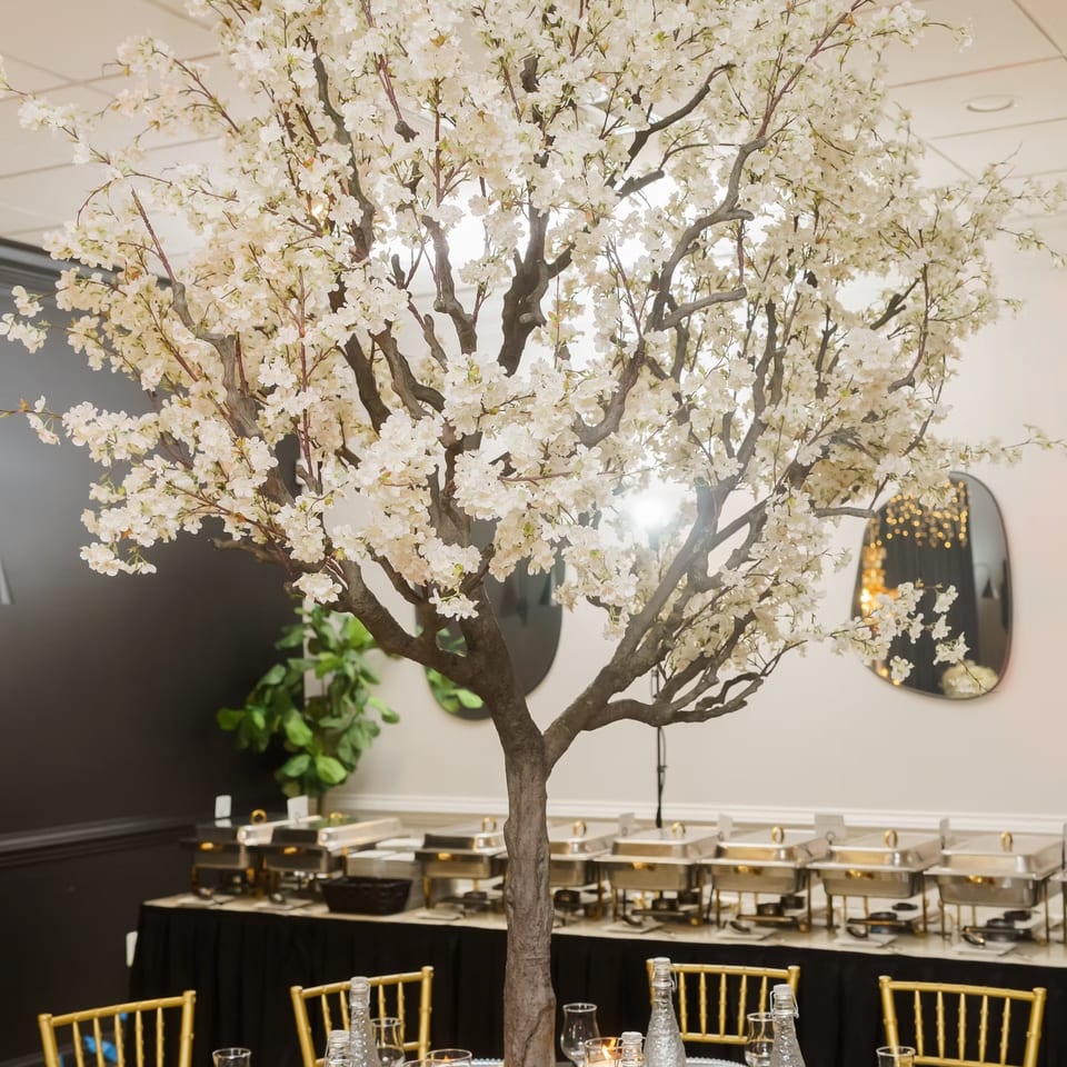 Elegant banquet dining room with a large white-blossomed tree centerpiece, candlelit table settings, gold chairs, and buffet stations.