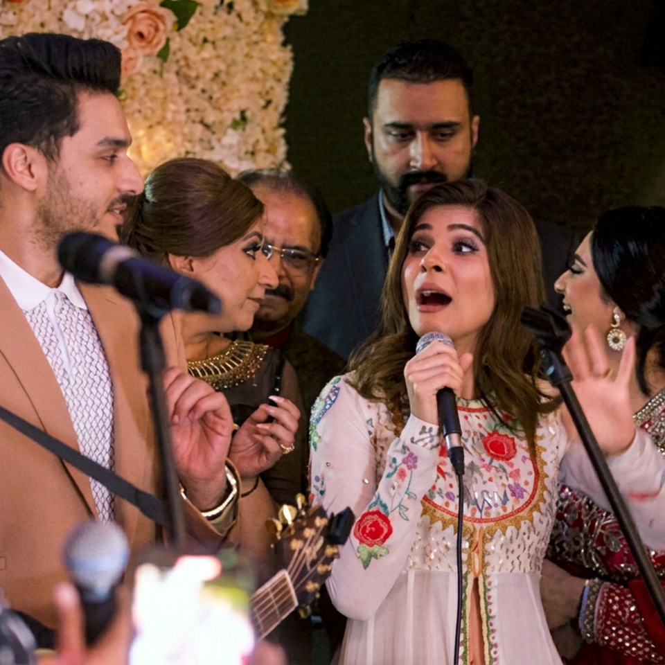 Woman singing into a microphone onstage with musicians and formally dressed guests gathered around her.