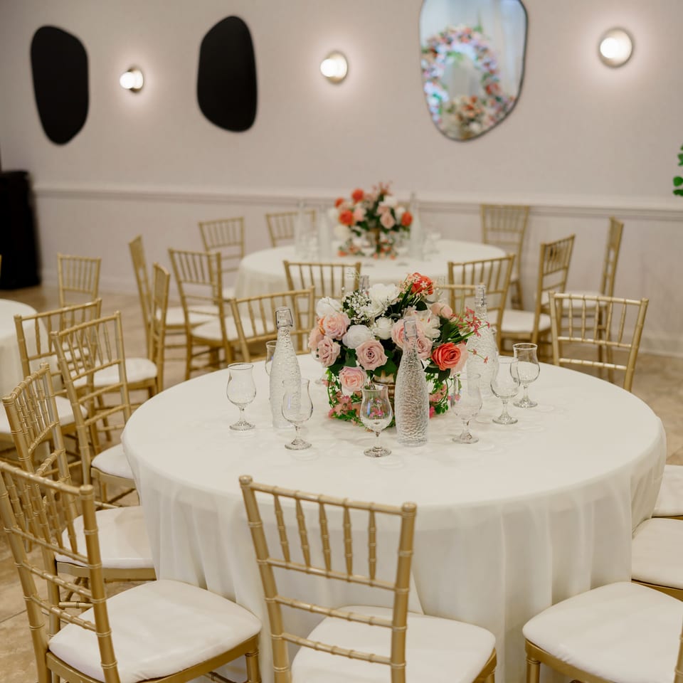 Elegant banquet dining room with round white-clothed tables, gold chairs, floral centerpieces, glassware, and decorative plants.