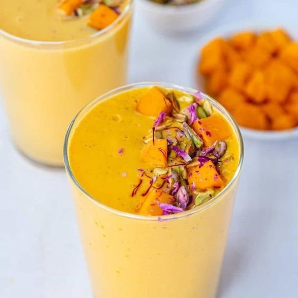 Mango smoothie in a glass topped with mango cubes, pistachios, rose petals, and saffron, with additional servings in the background.