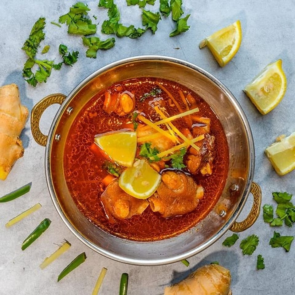 Overhead view of a spicy red curry with chicken, vegetables, lemon wedges, ginger, chilies, and cilantro in a metal serving dish.