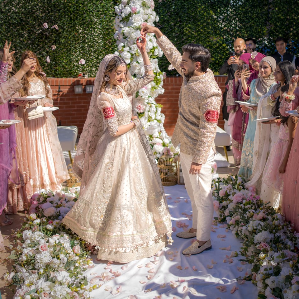 Bride and groom in ornate South Asian wedding attire dance amid flower arrangements while guests celebrate outdoors.