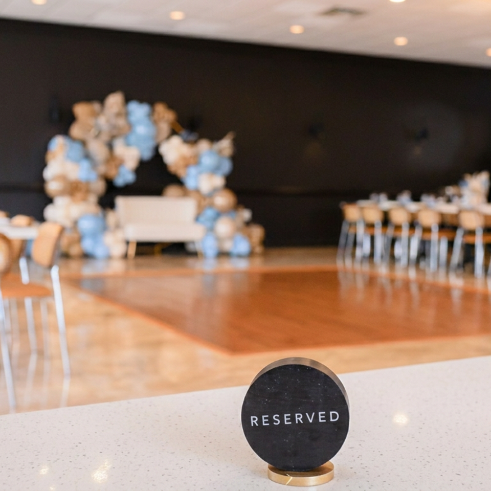 Reserved table sign in a decorated event hall with blue-and-white balloon arrangements, banquet tables, and chairs.