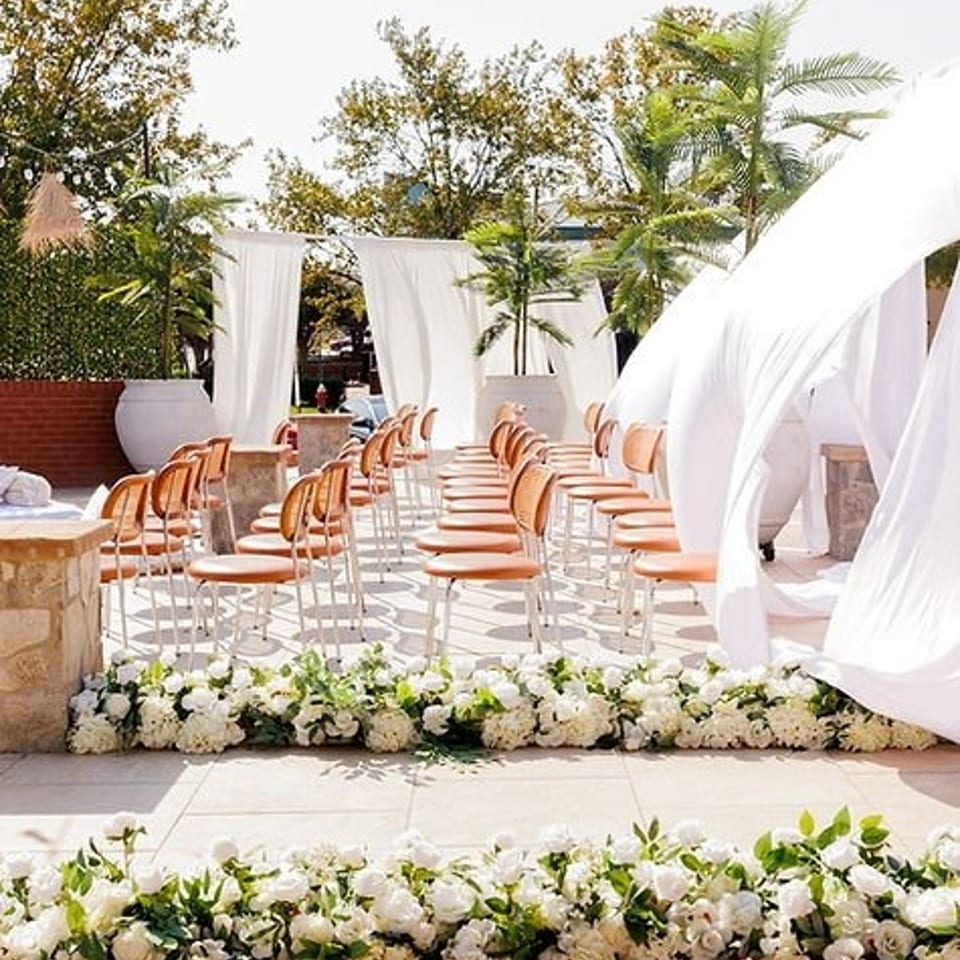 Elegant outdoor restaurant patio arranged for a ceremony, with white drapery, rows of peach chairs, tropical greenery, and white floral arrangements.