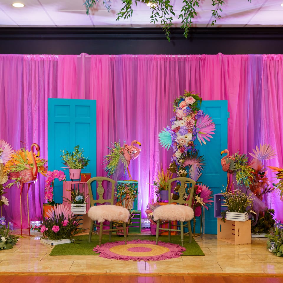 Colorful event backdrop with pink curtains, turquoise doors, floral arrangements, flamingo decorations, and two fur-covered chairs.