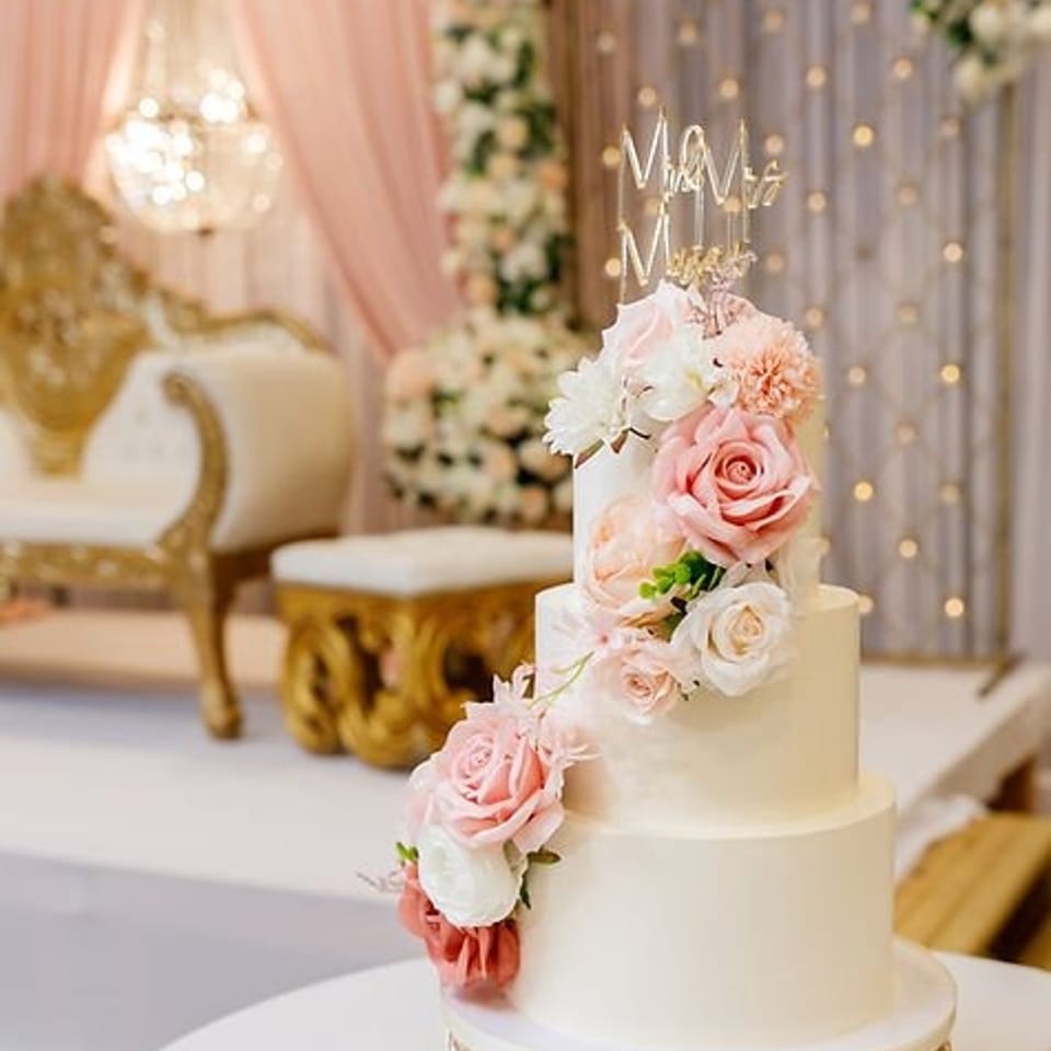 Elegant tiered wedding cake decorated with cascading pink and white roses, set in a softly lit floral wedding venue.