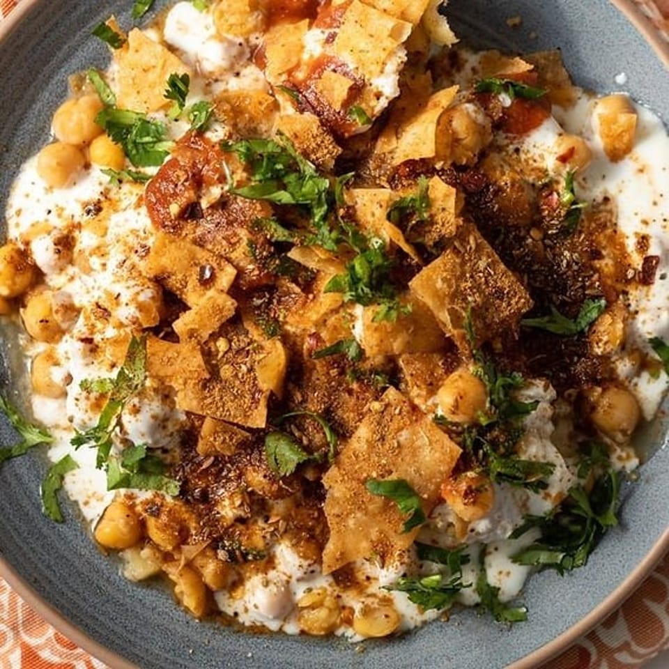 Overhead view of chickpeas, creamy yogurt, crispy pita pieces, spices, and fresh herbs served on a blue plate.