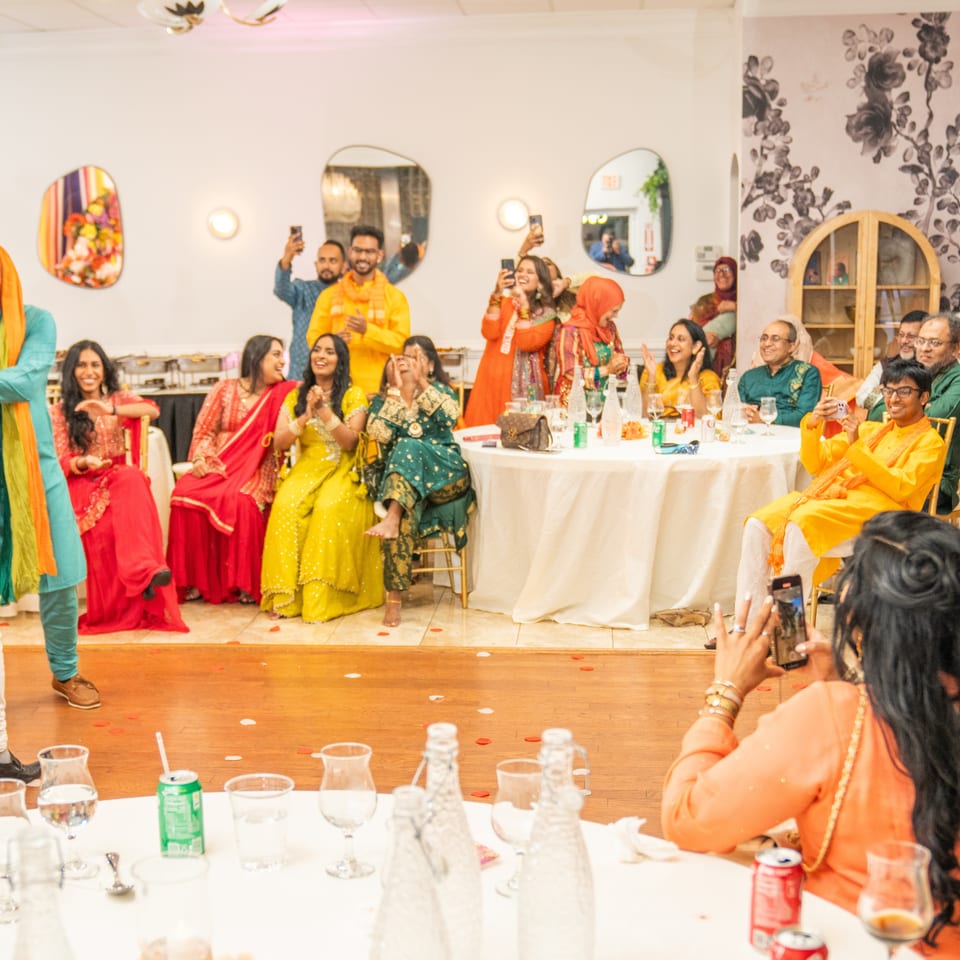 Guests in colorful South Asian attire laugh and celebrate at a restaurant banquet, with tables, drinks, and a photographer.