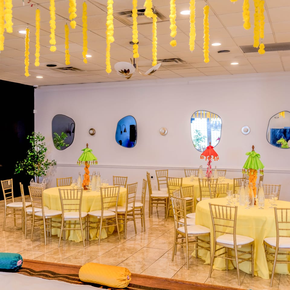 Elegant banquet hall with yellow-draped tables, gold chairs, colorful umbrella centerpieces, and hanging yellow floral decorations.