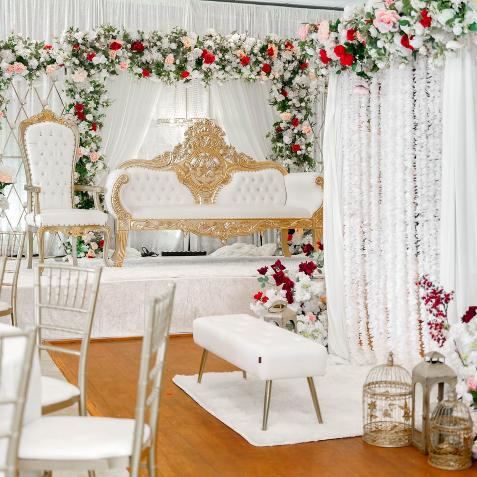 Ornate white and gold wedding stage with loveseat, throne chair, floral arches, draped curtains, and guest seating.