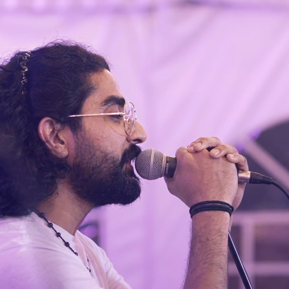 Bearded musician with long hair and glasses sings into a microphone under purple stage lighting.