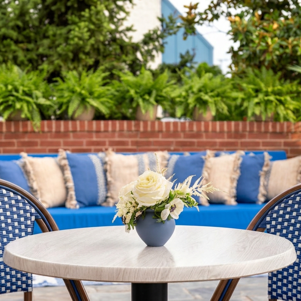 Outdoor restaurant patio with a round table, blue woven chairs, floral centerpiece, cushioned bench, brick wall, and lush plants.