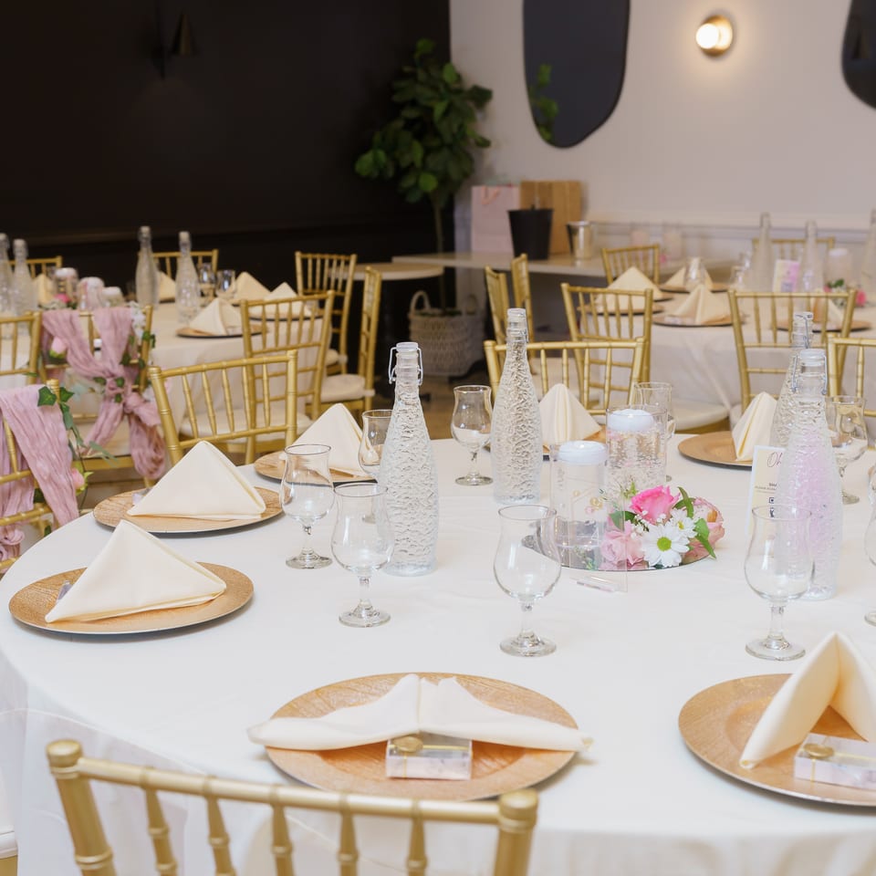 Elegant restaurant dining room with white tablecloths, gold chairs, folded napkins, glassware, floral centerpieces, and decorative water bottles.