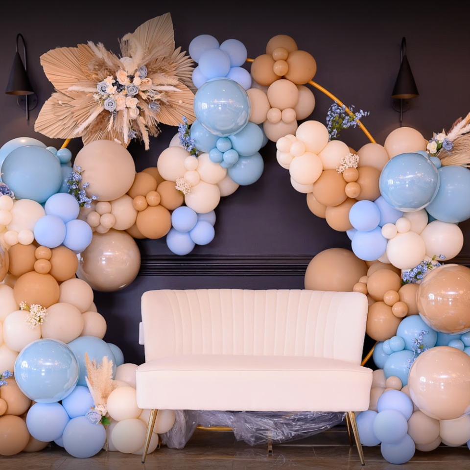 White loveseat framed by blue, peach, and cream balloon garlands, dried flowers, and decorative wall sconces.