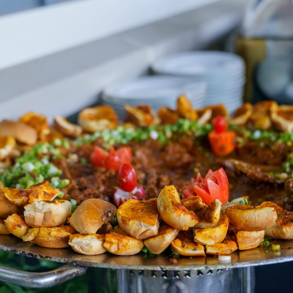 Large platter of grilled bread, roasted vegetables, chopped herbs, tomatoes, and savory meat prepared in a restaurant kitchen.